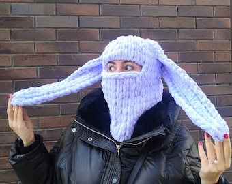 May include: A light purple, knitted balaclava with long, floppy bunny ears. The face covering has an opening for the eyes and mouth. The hat is made of a thick, soft yarn and is worn by a person wearing a black jacket.