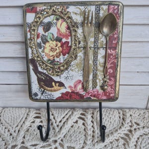Op de afbeelding: Een decoratieve wandhaak met een bloemenpatroon en een vogel. De haak is gemaakt van hout en heeft een gouden frame. Er zijn twee haken aan de onderkant van het stuk. De haak is versierd met een vork en een lepel.