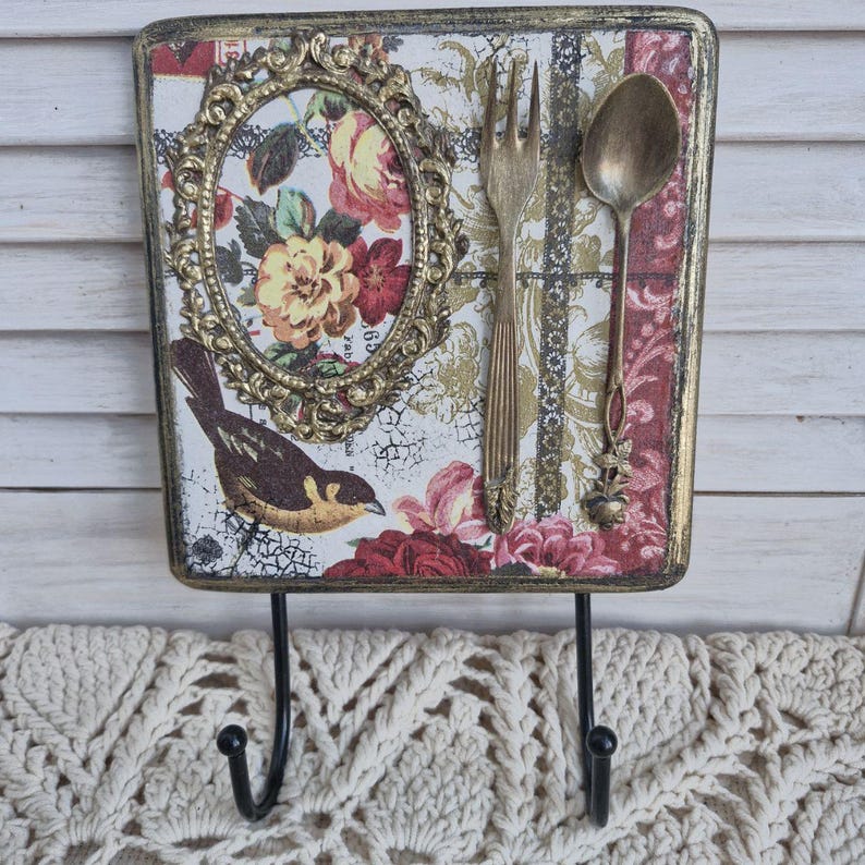 Op de afbeelding: Een decoratieve wandhaak met een bloemenpatroon en een vogel. De haak is gemaakt van hout en heeft een gouden frame. Er zijn twee haken aan de onderkant van het stuk. De haak is versierd met een vork en een lepel.