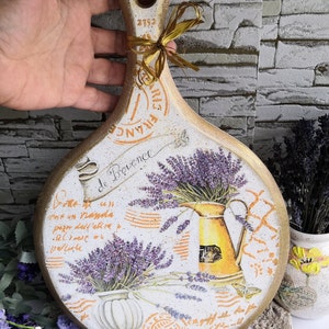 Pu&ograve; includere: Un tagliere rotondo in legno con manico, decorato con un tema di lavanda. Il tagliere presenta un design in stile vintage con fiori di lavanda, una brocca e un vaso decorativo. Il testo "Bode de Provence" &egrave; scritto sul tagliere.