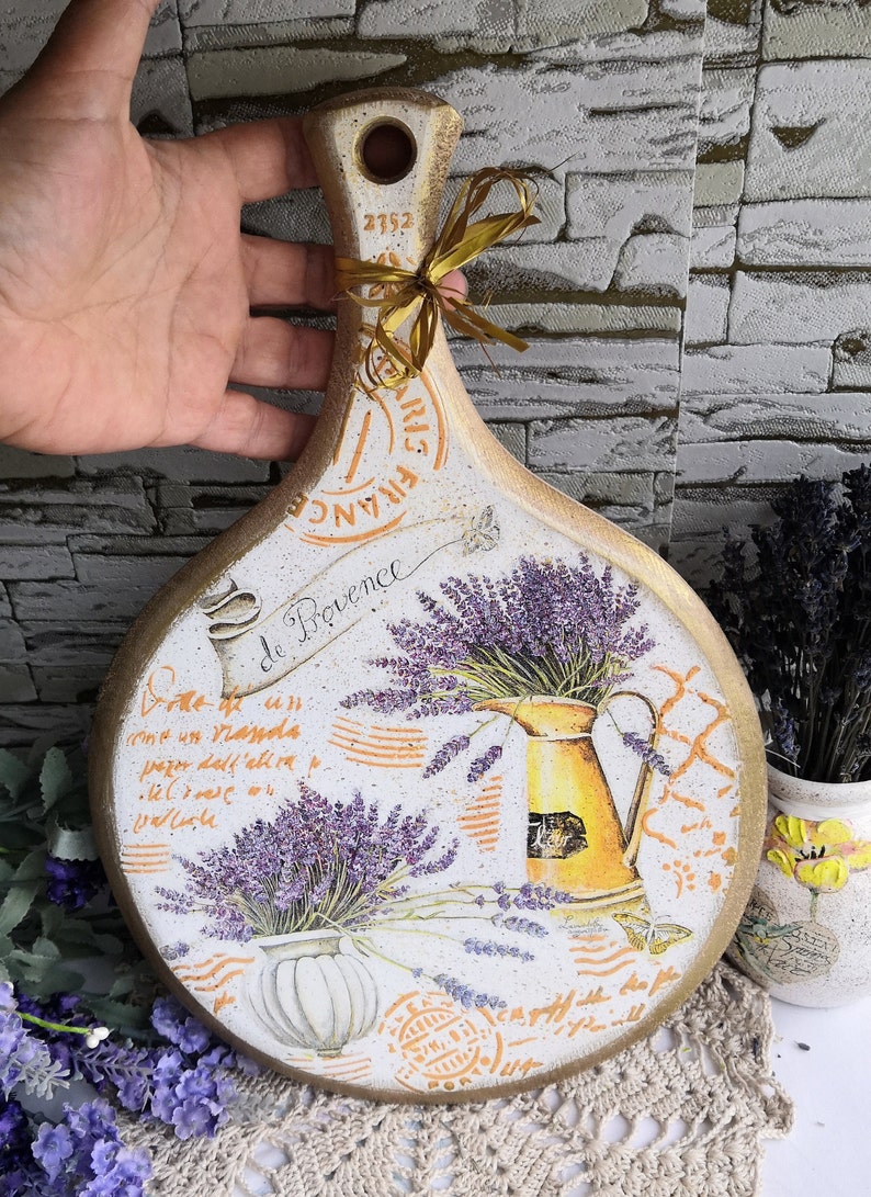 Pu&ograve; includere: Un tagliere rotondo in legno con manico, decorato con un tema di lavanda. Il tagliere presenta un design in stile vintage con fiori di lavanda, una brocca e un vaso decorativo. Il testo "Bode de Provence" &egrave; scritto sul tagliere.