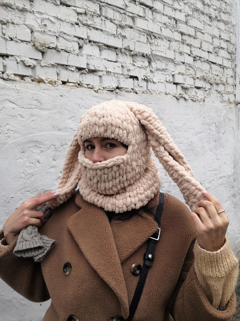 Puede incluir: Un pasamonta&ntilde;as beige tejido con largas orejas de conejo flexibles. La cobertura facial tiene una textura acanalada y se usa con un abrigo marr&oacute;n. El sombrero es un accesorio &uacute;nico hecho a mano.