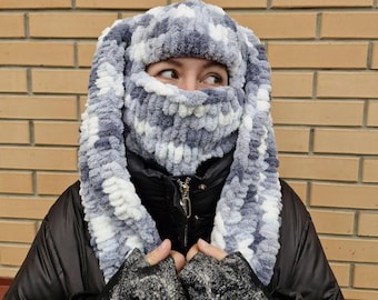 May include: A gray and white knitted balaclava with long ear flaps. The balaclava is made of a soft, fluffy yarn.