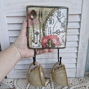 Op de afbeelding: Een decoratieve wandhaak met een vintage klok, een bloemenontwerp en sleutels. Twee beige keramische mokken hangen aan de haken.