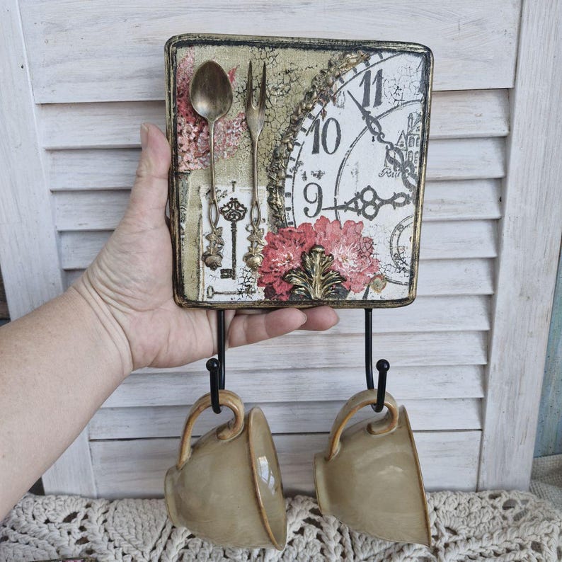 Op de afbeelding: Een decoratieve wandhaak met een vintage klok, een bloemenontwerp en sleutels. Twee beige keramische mokken hangen aan de haken.