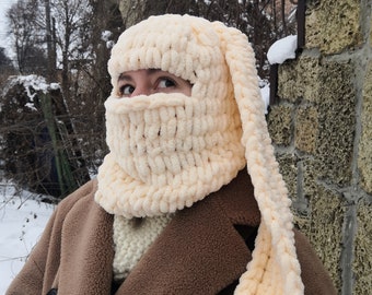 May include: A cream-colored knitted hat with long ear flaps. The hat is made of a soft, fluffy yarn and has a loose, slouchy fit.