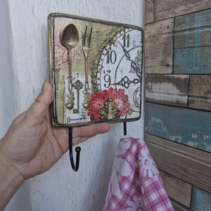 Op de afbeelding: Een decoratieve houten wandhaak met een vintage klok, bloemmotieven en een gouden lepel en vork. De haak heeft twee zwarte metalen haken die aan de onderkant hangen.