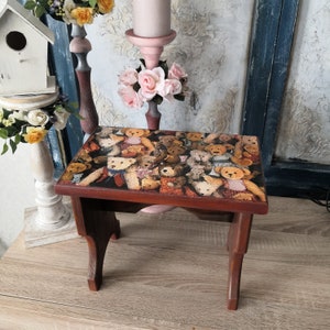 May include: A wooden stool with a brown finish and a top decorated with a collage of teddy bears. The stool has three legs and a rectangular top.