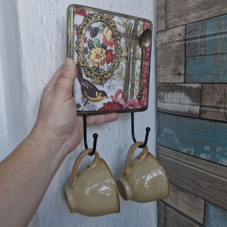 Op de afbeelding: Een decoratieve houten wandhaak met een bloemenpatroon en een vogel. De haak heeft twee metalen haken om items aan op te hangen. Twee beige keramische mokken hangen aan de haken.