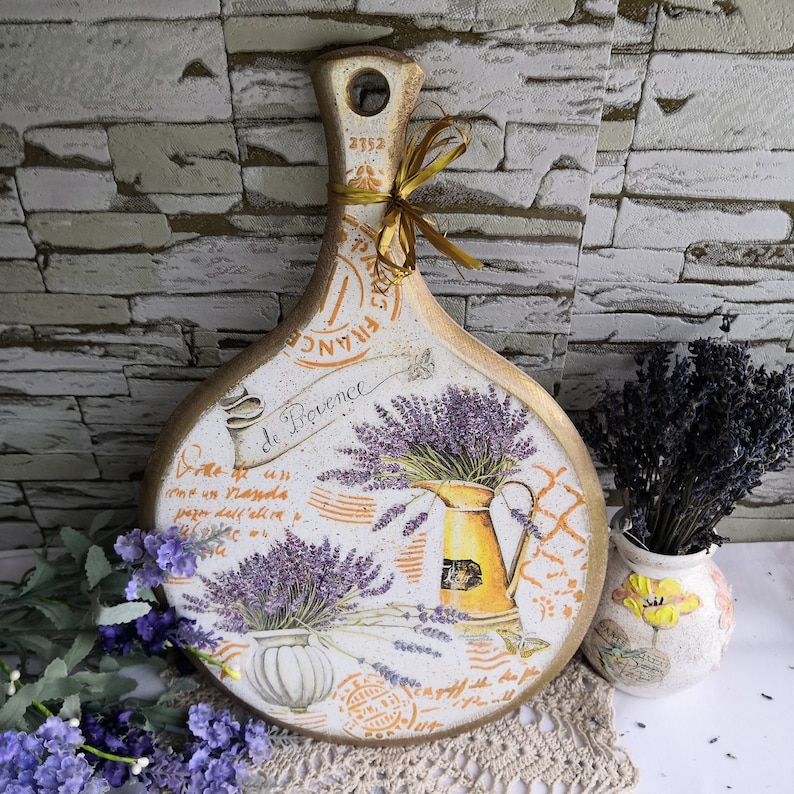Pu&ograve; includere: Un tagliere di legno bianco con un bordo dorato e un design di lavanda. Il tagliere ha il testo "Le Provence" e "France".