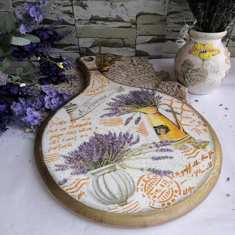 Pu&ograve; includere: Un tagliere rotondo in legno con un design dipinto di fiori di lavanda, una brocca e un vaso. Il tagliere ha un bordo dorato e le parole "de Provence" scritte su di esso.