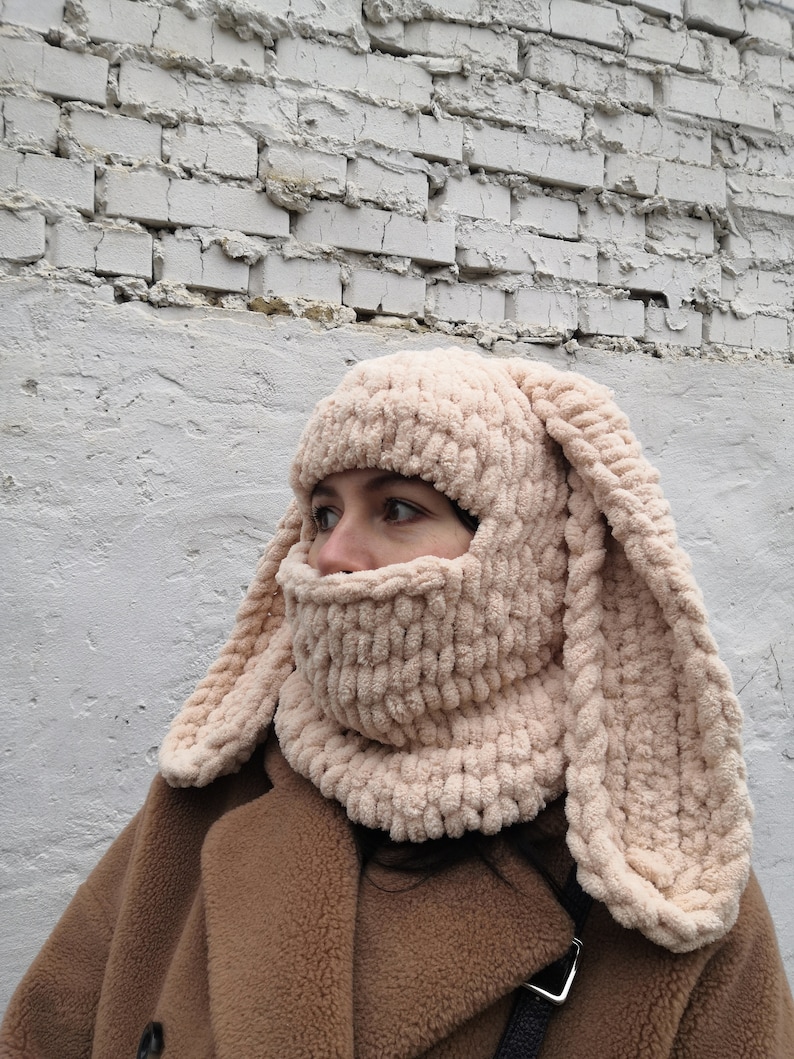 Puede incluir: Un pasamonta&ntilde;as beige tejido con largas orejas de conejo flexibles. La cobertura facial est&aacute; hecha de un material de punto suave y grueso. El pasamonta&ntilde;as se usa con un abrigo marr&oacute;n contra una pared de ladrillos blancos.