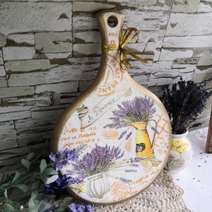 Pu&ograve; includere: Un tagliere rotondo in legno con manico, dipinto con un tema di lavanda. Il tagliere presenta un disegno floreale con fiori di lavanda, una brocca e il testo "de Provence".