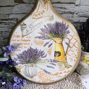 Pu&ograve; includere: Un tagliere rotondo in legno con un tema di lavanda. Il tagliere &egrave; decorato con un motivo floreale con fiori di lavanda, una brocca e un vaso. Il tagliere ha uno sfondo bianco con accenti arancioni e marroni. Il testo "Save de Provence" &egrave; scritto sul tagliere.