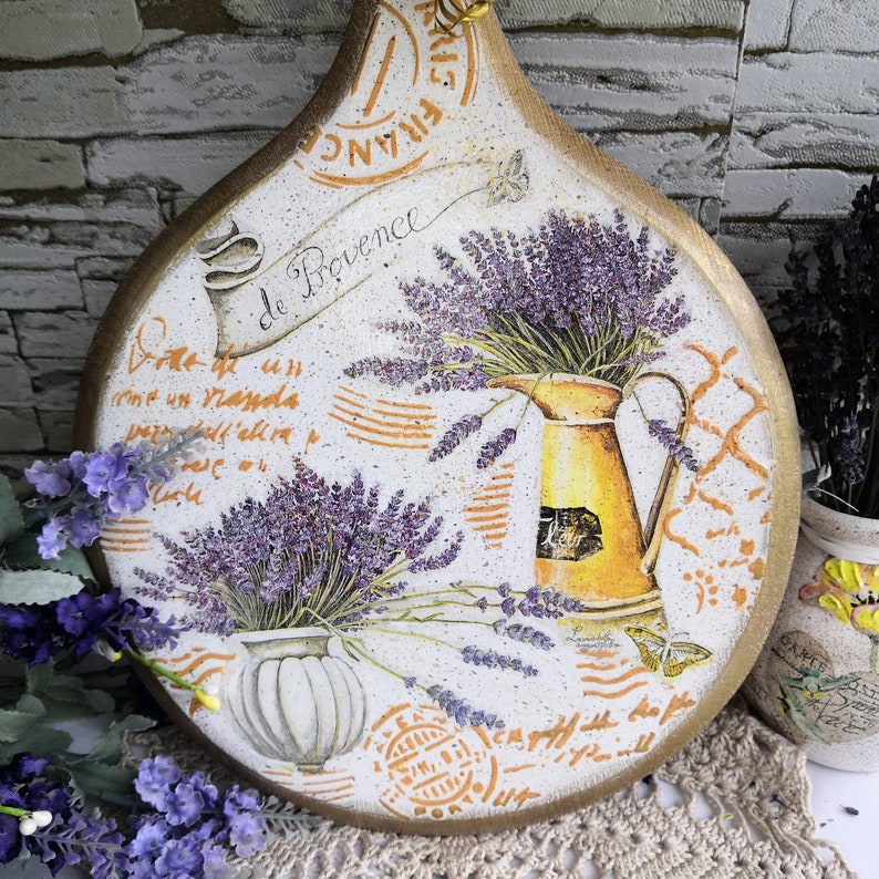 Pu&ograve; includere: Un tagliere rotondo in legno con un tema di lavanda. Il tagliere &egrave; decorato con un motivo floreale con fiori di lavanda, una brocca e un vaso. Il tagliere ha uno sfondo bianco con accenti arancioni e marroni. Il testo "Save de Provence" &egrave; scritto sul tagliere.