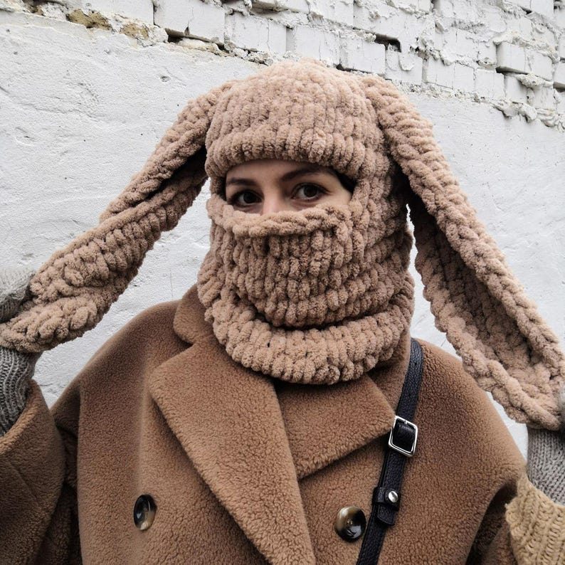 Puede incluir: Un pasamonta&ntilde;as tejido de color beige, con largas orejas de conejo ca&iacute;das. La cara est&aacute; cubierta por el gorro, y el material parece suave y afelpado. El pasamonta&ntilde;as se lleva con un abrigo marr&oacute;n.