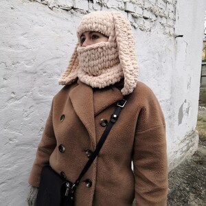 Puede incluir: Un pasamonta&ntilde;as beige de punto grueso con largas orejas de conejo ca&iacute;das. La cobertura facial est&aacute; integrada en el sombrero, creando un accesorio de invierno acogedor y caprichoso. La persona lleva un abrigo marr&oacute;n y un bolso negro.