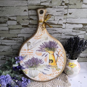 Pu&ograve; includere: Un tagliere in legno bianco e dorato con un design di lavanda e il testo "De Provence" e "France".