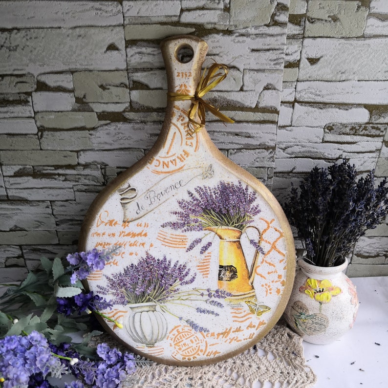 Pu&ograve; includere: Un tagliere in legno bianco e dorato con un design di lavanda e il testo "De Provence" e "France".