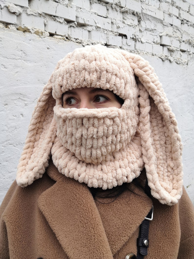 Puede incluir: Un pasamonta&ntilde;as beige tejido con largas orejas de conejo flexibles. La cobertura facial tiene una abertura para los ojos y la boca. El sombrero est&aacute; hecho de un material suave y afelpado. La persona lleva un abrigo marr&oacute;n.