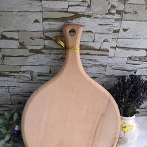 Pu&ograve; includere: Un tagliere rotondo in legno con un manico. Il tagliere &egrave; in legno chiaro e ha una finitura naturale. Il manico &egrave; attaccato alla parte superiore del tagliere e ha un piccolo foro al centro.