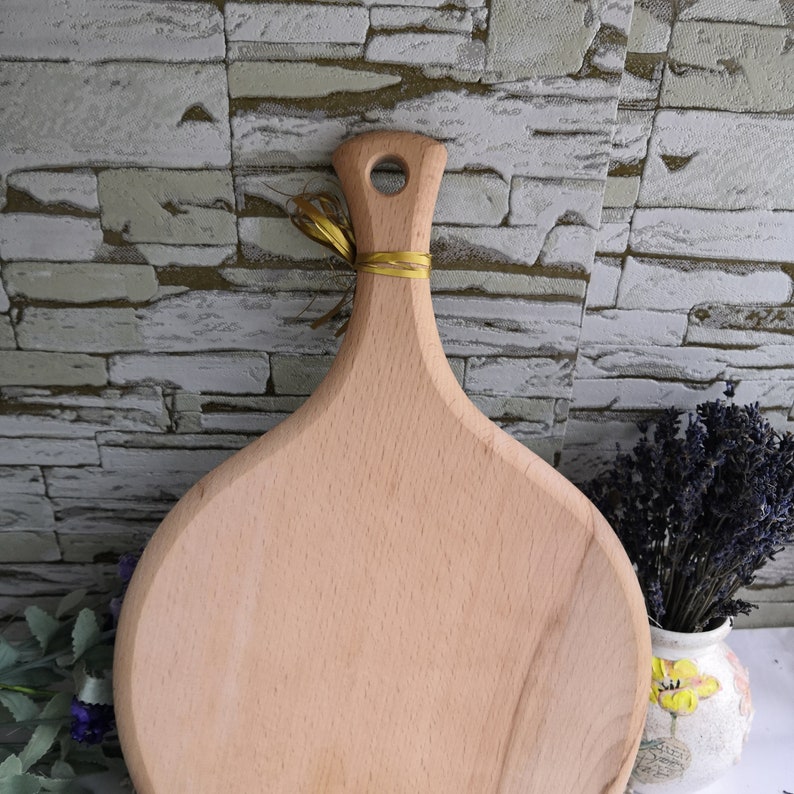 Pu&ograve; includere: Un tagliere rotondo in legno con un manico. Il tagliere &egrave; in legno chiaro e ha una finitura naturale. Il manico &egrave; attaccato alla parte superiore del tagliere e ha un piccolo foro al centro.