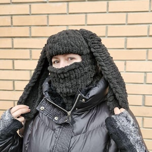Puede incluir: Un pasamonta&ntilde;as negro tejido con largas orejas de conejo flexibles. La cobertura facial tiene un dise&ntilde;o texturizado y acanalado. La persona lleva un abrigo de invierno negro y guantes grises. El fondo es una pared de ladrillos.