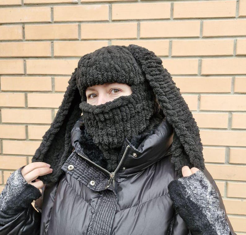 Puede incluir: Un pasamonta&ntilde;as negro tejido con largas orejas de conejo flexibles. La cobertura facial tiene un dise&ntilde;o texturizado y acanalado. La persona lleva un abrigo de invierno negro y guantes grises. El fondo es una pared de ladrillos.