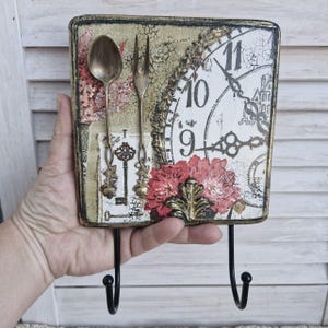 Op de afbeelding: Een decoratieve wandhaak met een verweerd, vintage-ge&iuml;nspireerd ontwerp. De haak heeft een klok met Romeinse cijfers, bloemmotieven en een sleutelgat. Twee haken zijn aan de onderkant van het stuk bevestigd.