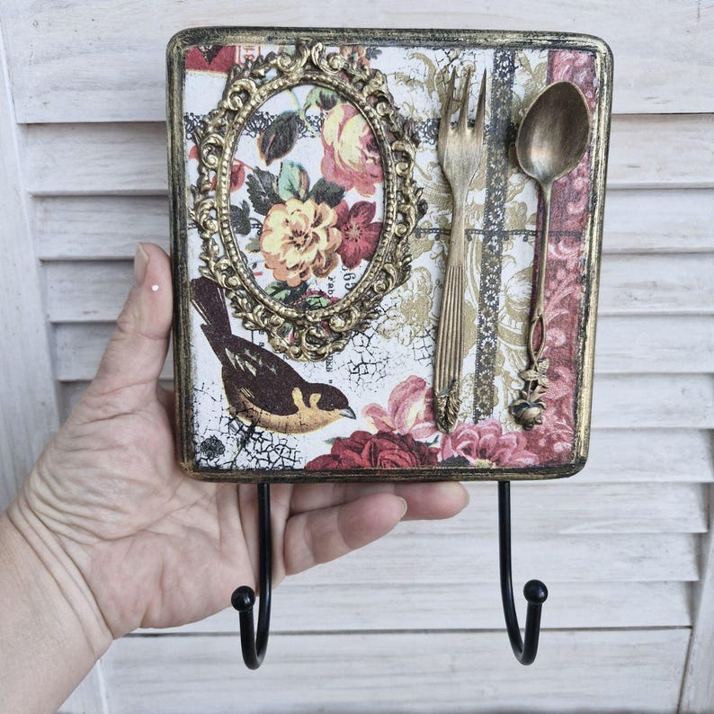 Op de afbeelding: Een decoratieve houten wandhaak met een bloemenmotief en een gouden frame. De haak heeft een vogel, een vork en een lepel. De haak is geschilderd in een verweerde witte en gouden afwerking.
