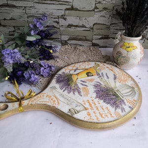 Pu&ograve; includere: Un tagliere rotondo in legno con manico, dipinto con un tema lavanda. Il tagliere ha un bordo dorato e presenta un design francese vintage con le parole "Douaiselle" e "Paris France".