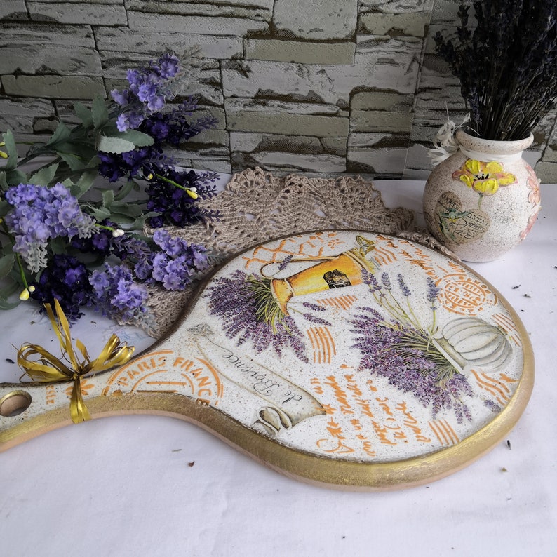 Pu&ograve; includere: Un tagliere rotondo in legno con manico, dipinto con un tema lavanda. Il tagliere ha un bordo dorato e presenta un design francese vintage con le parole "Douaiselle" e "Paris France".