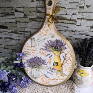 Pu&ograve; includere: Un tagliere di legno bianco con un design di lavanda e il testo "de Provence". Il tagliere ha un aspetto rustico e vintage ed &egrave; decorato con un motivo floreale e una brocca di lavanda.