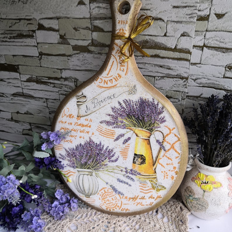Pu&ograve; includere: Un tagliere di legno bianco con un design di lavanda e il testo "de Provence". Il tagliere ha un aspetto rustico e vintage ed &egrave; decorato con un motivo floreale e una brocca di lavanda.