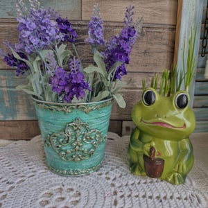 Może przedstawiać: Zielona ceramiczna doniczka w kształcie żaby z małym brązowym wiaderkiem w ręce. Żaba ma uśmiechniętą twarz i duże oczy. Żaba siedzi na białym powierzchni zrobionej na szydełku. Turkusowa metalowa doniczka z ozdobnym wzorem zawiera bukiet fioletowych kwiatów lawendy.