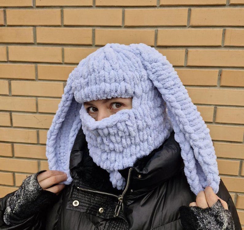 Puede incluir: Un pasamonta&ntilde;as tejido de color azul claro con largas orejas de conejo. El gorro cubre la cabeza y el cuello, con una abertura para los ojos y la boca. El gorro est&aacute; hecho de un hilo suave y texturizado y se usa sobre una chaqueta negra.