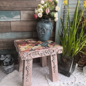 May include: A floral patterned stool with a distressed white finish and ornate details. The stool is sitting on a white lace tablecloth and has a vase of pink and white flowers behind it.
