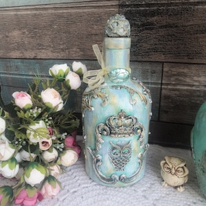 Puede incluir: Una botella de vidrio azul verdoso con un diseño de corona dorada y búho. La botella tiene un tapón decorativo y está decorada con detalles dorados. La botella está sobre una superficie blanca con un ramo de flores rosas y blancas.
