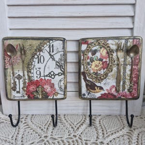 Op de afbeelding: Twee decoratieve wandhaken met een vintage ontwerp. Elke haak heeft een vierkante houten plaat met een ander ontwerp. E&eacute;n plaat heeft een klok met Romeinse cijfers en een bloemenmotief. De andere plaat heeft een vogel, een bloemenmotief en een gouden ovale lijst. Beide platen hebben een gouden rand en een zwarte metalen haak.
