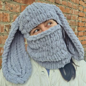 Puede incluir: Un gorro de conejo tejido de color gris claro con orejas largas y ca&iacute;das y una cobertura facial a juego. El gorro y la cobertura facial est&aacute;n hechos de un material suave y afelpado. La persona lleva una chaqueta blanca.