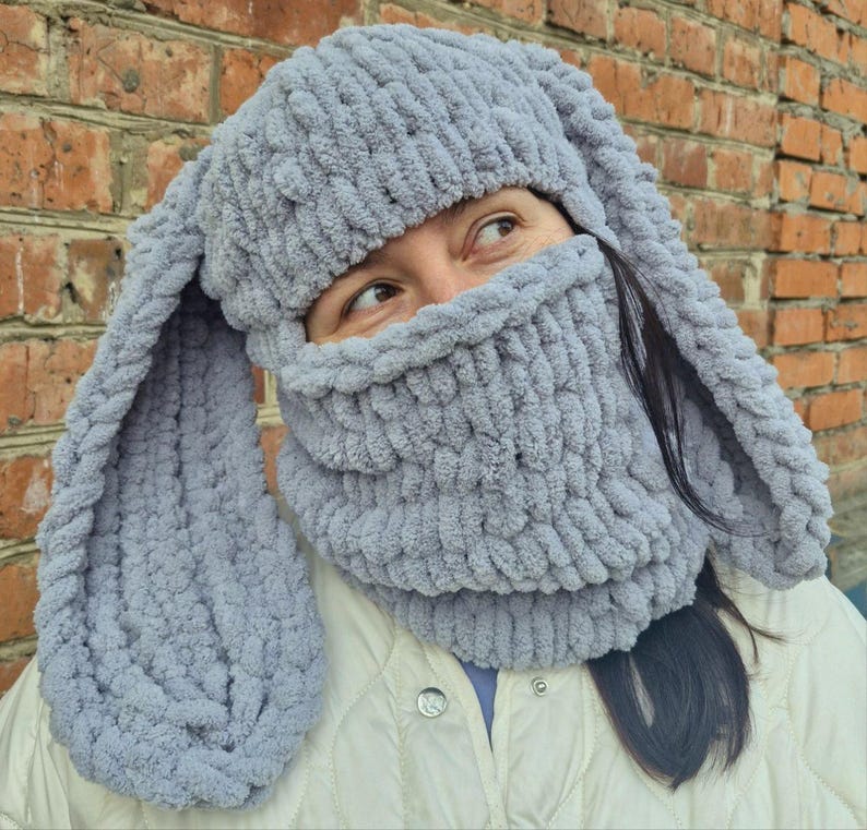 Puede incluir: Un gorro de conejo tejido de color gris claro con orejas largas y ca&iacute;das y una cobertura facial a juego. El gorro y la cobertura facial est&aacute;n hechos de un material suave y afelpado. La persona lleva una chaqueta blanca.