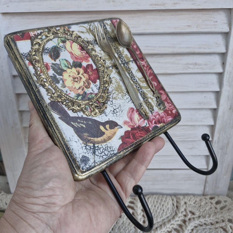Op de afbeelding: Een decoratieve wandhaak met een bloemenpatroon en een vogel. De haak is gemaakt van hout en heeft een gouden afwerking. Hij heeft twee zwarte metalen haken.