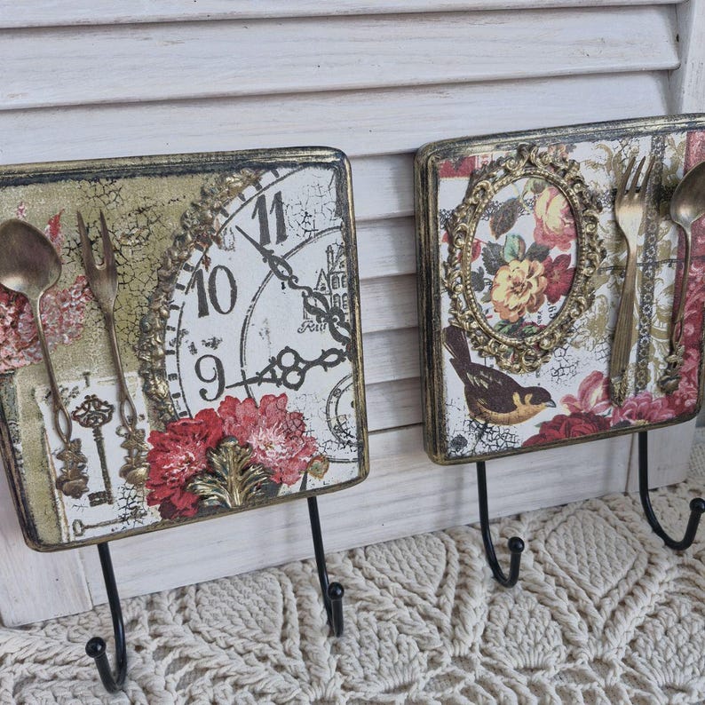Op de afbeelding: Twee decoratieve wandhaken met een vintage uitstraling. Elke haak heeft een vierkante houten plaat met een ander ontwerp. E&eacute;n plaat heeft een klok met Romeinse cijfers en een sleutelgat, terwijl de andere een vogel en een bloemenmotief heeft. Beide platen hebben een verweerde, antieke uitstraling en zijn versierd met gouden accenten.