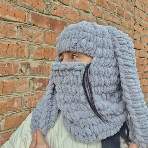 Puede incluir: Un gorro de conejo tejido de color gris claro con orejas largas y ca&iacute;das y una cobertura facial. El gorro est&aacute; hecho de un hilo suave y grueso y est&aacute; dise&ntilde;ado para cubrir la cabeza, el cuello y parte de la cara. El fondo es una pared de ladrillos.