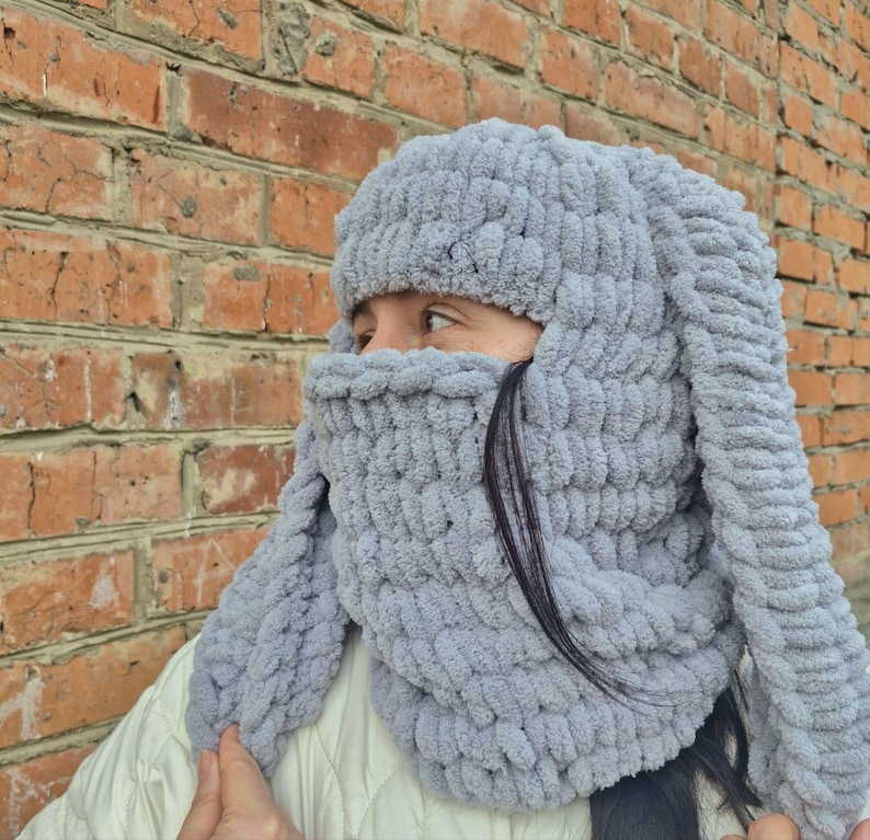 Puede incluir: Un gorro de conejo tejido de color gris claro con orejas largas y ca&iacute;das y una cobertura facial. El gorro est&aacute; hecho de un hilo suave y grueso y est&aacute; dise&ntilde;ado para cubrir la cabeza, el cuello y parte de la cara. El fondo es una pared de ladrillos.