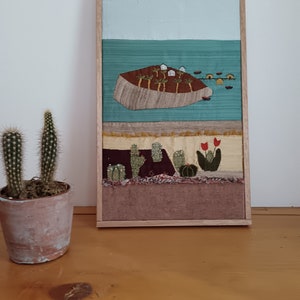 Puede incluir: Una obra de arte textil enmarcada que representa una escena de isla. La obra presenta una isla marrón con palmeras, edificios blancos y pequeños botes en un mar turquesa. El marco es de color marrón claro y un cactus en maceta está en primer plano.