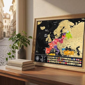 Op de afbeelding: Een ingelijste kraskaart van Europa met een zwarte achtergrond en goudkleurige landmassa's. Specifieke landen zijn gemarkeerd in levendige kleuren en onderaan staat een rij nationale vlaggen. Een kompasroos is zichtbaar in de hoek.