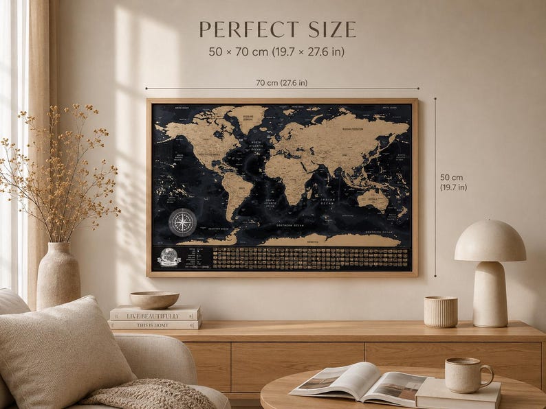 May include: A rectangular scratch-off world map in a light wood frame with black oceans and gold continents. The map features a compass rose and icons at the bottom. Text reads PERFECT SIZE 50 x 70 cm (19.7 x 27.6 in) with dimension lines.