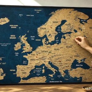 Op de afbeelding: Een ingelijste kraswereldkaart van Europa met een diepblauwe oceaanachtergrond en goudkleurige landmassa's. De kaart bevat gedrukte landnamen, grote steden en de tekst "EUROPE" en "TRAVEL STORY" met een wereldbolpictogram in de hoek.