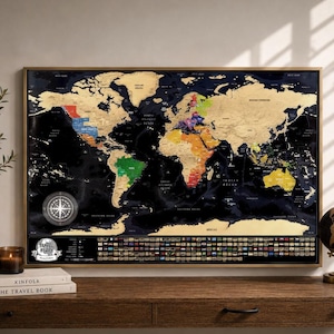 May include: A framed world map with a scratch-off gold foil layer, revealing colourful countries beneath. The map is displayed on a wooden desk, alongside a globe, books, and a decorative vase. The map measures approximately 91 cm wide and 61 cm tall.